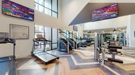 This expansive fitness center is designed to deliver a premium workout experience through a thoughtful combination of architectural flair and comprehensive equipment offerings. The space is immediately distinguished by its striking chevron-patterned luxury vinyl flooring, rendered in a mix of warm and cool tones that adds a dynamic visual energy underfoot.

Soaring double-height ceilings open the space dramatically, enhanced by an abundance of natural light streaming through large, floor-to-ceiling windows that offer views of an adjacent outdoor amenity area. Multiple ceiling fans are strategically distributed throughout to maintain airflow and comfort during workouts.

The cardio zone is well-appointed with a row of treadmills and stair climbers positioned along the window wall, each unit accompanied by an individual screen for entertainment. Multiple wall-mounted flat-screen televisions are placed throughout the facility, ensuring visibility from virtually every area of the gym.

Full-length mirrors line key walls, visually expanding the space and allowing users to monitor their form. The strength training area is generously outfitted with a multi-station cable machine, squat racks, and a complete set of free weights on tiered dumbbell racks. A beverage cooler and open storage cubbies near the entrance provide added convenience. Glass-paneled doors create a seamless, light-filled transition between zones.