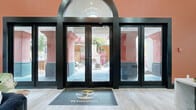 This single-floor residential clubhouse and leasing office for SZ Residential makes an immediate impression with its striking arched entry — framed by bold black-trimmed glass doors and floor-to-ceiling windows that offer a clear sightline to a beautifully landscaped outdoor courtyard beyond. A branded welcome mat anchors the entryway, setting a polished tone from the moment of arrival.

Inside, the space is divided into two distinct yet cohesive zones. The lounge area is appointed with plush, blush-pink curved accent chairs accented with gold bases, arranged around a marble-topped circular coffee table atop a sage green area rug. Dark emerald green velvet chairs and café-style bistro tables provide additional seating, while tall black-trimmed windows flood the room with natural light. A pendant chandelier with cascading greenery adds a lush, botanical touch overhead, and bold black crown molding throughout lends dramatic architectural contrast to the crisp white walls.

The second zone features soaring ceilings and a dramatic floor-to-ceiling geometric wood-paneled fireplace wall with a mounted television. A deep navy tufted sofa anchors this gathering space, complemented by olive green accent chairs. Adjacent to the lounge is a sleek bar-style welcome counter finished in marble and stone tile, backed by rich burgundy subway tile and a glowing neon sign reading "Welcome to the Good Life," completing this vibrant and welcoming community hub.