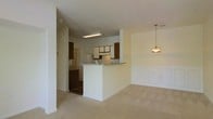 This single-floor residence features an open layout that connects the kitchen and living areas, creating a cohesive and functional living space. The kitchen is equipped with a mix of white and dark wood cabinetry, offering ample storage. A white peninsula extends outward, providing additional counter space and serving as a natural divider between the kitchen and the adjacent living area. Overhead, a fluorescent light fixture illuminates the kitchen, while a classic pendant light hangs above the dining area, adding a warm, decorative touch. The living area is finished with neutral beige carpeting, contributing to a soft and comfortable atmosphere. A notable design feature is the wainscoting along the lower portion of the living area walls, adding architectural interest and a traditional aesthetic. The overall color palette consists of warm whites and beiges, creating a bright and welcoming environment. A hallway visible in the background suggests additional rooms beyond the main living space, and the layout flows efficiently throughout the unit.