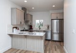 Located at 6328 W State St in Boise, Idaho, this two-story property showcases a clean, contemporary design throughout. The main level features an expansive open-plan living and kitchen area with warm hardwood flooring and recessed lighting that creates a bright, welcoming atmosphere. The kitchen is thoughtfully appointed with light wood cabinetry, white quartz countertops, a subway tile backsplash, stainless steel appliances including a gas range and refrigerator, and a matte black faucet over an undermount sink. A peninsula provides additional counter space and casual seating. A convenient full bathroom is also accessible on the main level. Upstairs, carpeted bedrooms offer a comfortable retreat, with windows framing views of the surrounding landscape, including open green spaces and mature trees. The bathrooms are finished with neutral tile flooring, wood-toned vanities with white quartz tops, and matte black hardware — one featuring a glass-enclosed walk-in shower with white subway tile, and the other a shower-tub combo. A carpeted staircase with a clean white handrail connects the two levels. Throughout the property, a consistent palette of whites, natural wood tones, and matte black accents lends a cohesive, modern aesthetic.
