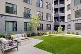 Located at 8448 Delmar Blvd in University City, Missouri, this thoughtfully designed outdoor courtyard serves as a central amenity space nestled within a modern multi-story building. The courtyard is defined by its contemporary architectural aesthetic, with the surrounding structure showcasing a combination of charcoal-toned brick and light stucco facades, floor-to-ceiling windows, and private balconies overlooking the space below.

At ground level, expansive stretches of vibrant artificial turf provide a clean, low-maintenance lawn area ideal for recreation or relaxation. Wide, light-toned concrete paver pathways weave throughout the courtyard, connecting various zones seamlessly. A dedicated seating area features a stone fire pit surrounded by comfortable outdoor furniture, creating an inviting gathering spot for residents.

Mature landscaping beds line the perimeter of the courtyard, planted with a diverse mix of evergreen shrubs, ornamental grasses, young deciduous trees, and ground-cover plantings set in mulched beds—adding natural texture and seasonal color. Glass entry doors with metal frames provide interior access directly from the courtyard. The open-air design allows ample natural light to filter into the space throughout the day, while the building's enclosing wings offer a sense of privacy and shelter, making this courtyard a serene and functional outdoor retreat.
