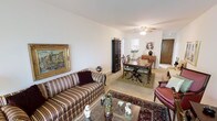 This single-floor residence presents an open-plan living and dining area that functions as the central hub of the home. The living space is furnished with a classic striped sofa and a leather armchair, arranged around a glass-top coffee table adorned with decorative accents. A richly patterned area rug anchors the seating arrangement, adding warmth and visual depth to the neutral-toned flooring beneath. A large gilt-framed painting on the wall contributes an elegant, traditional character to the room.

The dining area flows naturally from the living space and features a dark wood dining table paired with upholstered chairs, set atop a complementary area rug. A tall dark wood cabinet provides storage and serves as a refined design element. The ceiling fan overhead offers both function and a classic aesthetic touch.

Beyond the dining area, a glimpse into what appears to be a kitchen or additional room is visible, suggesting a practical and connected layout. Natural light filters through the space, enhancing the warm, inviting ambiance. Overall, the home conveys a traditional, well-appointed character with cohesive décor throughout.