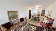 This single-floor residence presents an open-plan living and dining area that functions as the central hub of the home. The living space is furnished with a classic striped sofa and a leather armchair, arranged around a glass-top coffee table adorned with decorative accents. A richly patterned area rug anchors the seating arrangement, adding warmth and visual depth to the neutral-toned flooring beneath. A large gilt-framed painting on the wall contributes an elegant, traditional character to the room.

The dining area flows naturally from the living space and features a dark wood dining table paired with upholstered chairs, set atop a complementary area rug. A tall dark wood cabinet provides storage and serves as a refined design element. The ceiling fan overhead offers both function and a classic aesthetic touch.

Beyond the dining area, a glimpse into what appears to be a kitchen or additional room is visible, suggesting a practical and connected layout. Natural light filters through the space, enhancing the warm, inviting ambiance. Overall, the home conveys a traditional, well-appointed character with cohesive décor throughout.