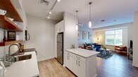 Located at 240 Sidney St in Cambridge, MA, this thoughtfully designed one-bedroom, one-bathroom residence offers a smart and efficient layout that maximizes every inch of its single-floor footprint. The open-concept kitchen and living area serve as the heart of the home, featuring a central island with white cabinetry, quartz countertops, a mosaic tile backsplash, stainless steel appliances including a refrigerator, dishwasher, range, and built-in microwave, plus open wood shelving that adds warmth and character. Pendant lighting over the island and track lighting enhance the clean, modern aesthetic. Wood-look flooring flows throughout the main living areas, complementing the bright, neutral palette. The living room benefits from large windows that frame green outdoor views and fill the space with natural light, while a bold geometric accent wall adds a distinctive design touch. The bedroom is a calm retreat with a generous window, plush carpeting, and a dedicated walk-in closet with built-in shelving. The bathroom is finished with tile flooring, a white vanity, and a glass-enclosed shower. A dedicated laundry area adds everyday convenience, completing this well-appointed Cambridge residence.