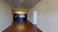 This single-story residence offers an open-concept layout where the kitchen and living areas flow seamlessly together, creating a cohesive and functional living space. Rich hardwood flooring extends throughout the main areas, providing a warm and inviting foundation.

The kitchen is a standout feature, outfitted with dark espresso cabinetry that extends to the ceiling, complemented by granite-style countertops. A built-in wine rack adds a distinctive design detail above the upper cabinets. Black appliances include a refrigerator, range, microwave, and dishwasher. A center island with matching countertop offers additional prep space and casual seating. Pendant lighting and track lighting illuminate the kitchen effectively.

The living area is generously proportioned, benefiting from natural light through windows near the front entry, which also features a ceiling fan. Two access doors are visible from this space, offering connectivity to other areas of the unit.

The bedroom features plush carpeting, a wide multi-panel window that brings in natural light, and a ceiling fan for comfort. An adjoining hallway leads to the bathroom, which includes a dark-cabinet vanity with granite-style countertop, a large mirror, and a tub-shower combination, with continued hardwood flooring at the transition point.
