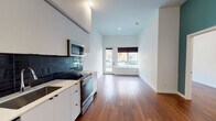 This single-floor unit offers a thoughtfully designed open-concept layout that seamlessly integrates the kitchen and living area into one cohesive space. The kitchen is a standout feature, outfitted with flat-front white cabinetry extending to the ceiling for generous storage, a striking dark subway tile backsplash, white countertops, and a full suite of stainless steel appliances including a refrigerator, dishwasher, range, and over-the-range microwave. Warm-toned hardwood flooring runs throughout the main living areas, grounding the bright, white walls and creating a welcoming contrast. The living area is bathed in natural light from a full glass door that opens to the outdoors, enhancing the sense of openness in the space. The bathroom is cleanly finished with white tile, a single-sink vanity with white cabinetry, a large wall mirror, and a tub-shower combination. A hallway leads to dedicated closet areas, providing practical storage solutions. Recessed lighting and tall ceilings contribute to an airy, modern ambiance throughout. The overall design is clean, minimalist, and move-in ready.
