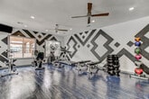 This fitness center makes an immediate visual impact with its eye-catching black-and-white geometric mural that spans the entirety of its walls, featuring large diamond and chevron patterns that lend the space a modern, energetic character. The dark hardwood-style flooring provides a sharp contrast to the bright white walls, reinforcing the bold aesthetic throughout.

The space is thoughtfully divided into dedicated cardio and strength training zones. The cardio area is well-stocked with a treadmill, elliptical trainer, stationary bike, and a cycle bike, all positioned near large windows that let in generous natural light and offer views of the surrounding outdoor greenery. A ceiling fan overhead ensures comfortable air circulation during intense workouts.

The strength training section features a multi-station cable machine, a weight bench, and a dumbbell rack, providing users with a comprehensive range of resistance training options. Recessed lighting throughout the ceiling supplements natural light and ensures the space remains bright and functional at all times.

The room's single-floor layout maximizes usability, keeping all equipment easily accessible. The combination of vibrant design, diverse equipment offerings, and a well-lit, ventilated environment makes this fitness center both visually stimulating and highly functional for a complete workout experience.