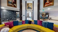 This striking single-floor lobby space makes an immediate impression with its soaring double-height ceilings and an eclectic, curated aesthetic that blends bold color with artistic expression. At the heart of the space sits a dramatic curved yellow sofa adorned with richly colored throw pillows in jewel tones of teal, magenta, and plum, anchored by a plush area rug and a round, multicolored ottoman — creating an inviting central lounge area. Dark structural columns contrast sharply against the bright white walls, adding architectural rhythm throughout the open floor plan. One wall features an expansive gallery display of mixed-media artwork in varied frames, ranging from classic portrait reproductions to contemporary prints, lending the space a museum-like quality. An elaborate chalk mural adorns a nearby wall, adding a hand-crafted, urban artistic touch. Decorative textured panels provide visual separation between zones while maintaining an open feel. A convenience snack bar with bar-height seating and a stocked refrigerator is positioned toward one end, offering a casual refreshment area. Lush potted indoor plants, colorful area rugs, and thoughtfully placed seating vignettes complete the design, resulting in a dynamic, welcoming communal environment that balances creativity with comfort.