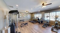 Located at 720 Curtis Pkwy SE in Calhoun, Georgia, this fitness center offers a comprehensive workout environment within a single, efficiently organized space. The room features warm hardwood-style flooring throughout, complemented by neutral sage-green walls and a clean white ceiling with recessed lighting and dual ceiling fans that ensure consistent airflow and comfort during exercise.

The facility is thoughtfully equipped with a diverse range of workout options. Cardio equipment includes multiple treadmills, elliptical trainers, and a stationary bike, positioned to take advantage of natural light from the windows fitted with blinds. A cable and pulley strength machine anchors the strength-training area, accompanied by a dedicated free weight rack stocked with a full set of dumbbells and adjustable benches for various exercises. Additional accessories include medicine balls, stability balls, and resistance bands neatly organized on a storage rack, supporting a wide range of functional fitness routines.

A wall-mounted flat-screen television adds an entertainment element to the space. Colorful framed artwork lines one wall, adding a vibrant, motivating atmosphere. The facility also includes a closet and access door, suggesting additional storage or utility space. Overall, this fitness center provides a well-rounded, practical environment suited to a full spectrum of workout needs.