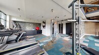 Located at 222 West Blvd in Charlotte, NC, this modern two-floor fitness center is thoughtfully designed to support a comprehensive workout experience. The main floor features an open layout with a vibrant patchwork carpet in shades of teal, gray, and tan, adding a contemporary aesthetic to the space. This level is equipped with a robust selection of cardio machines, including treadmills and ellipticals, alongside cable machines, free weights, weight racks, kettlebells, and a punching bag. A decorative mosaic tile accent wall and bold graphic wall coverings contribute to the dynamic atmosphere, while track lighting ensures the space is well-lit throughout. Large windows allow natural light to filter in, offering views of the surrounding Charlotte streetscape. A striking spiral staircase connects the main floor to the upper level, which features a more intimate setting with plush gray carpet, full-length mirrors, and a warmly lit geometric patterned accent wall. This upper area is furnished with weight benches and ab equipment, ideal for focused strength training. Industrial-style metal railings frame the mezzanine, completing the modern design. Together, the two floors create a well-rounded, stylish fitness facility suited for a variety of training needs.