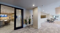 This thoughtfully designed single-floor commercial space combines contemporary aesthetics with functional versatility. The entry corridor makes an immediate impression with a bold dark brick-tile accent wall contrasted against light tile flooring, setting a sophisticated tone throughout. A central hallway lined with patterned carpet connects the various zones, flanked by sleek black-framed glass partition walls that enclose private conference and meeting rooms. These glass-walled rooms offer visibility while maintaining acoustic separation, with one featuring a large display screen and executive seating for formal presentations.

The open collaborative area serves as the heart of the space, featuring a coffered ceiling with decorative gold-toned beams and globe pendant lighting above a communal workstation island. Neutral carpet, recessed lighting, and large floor-to-ceiling windows flood the area with natural light and panoramic outdoor views. Comfortable lounge seating is arranged throughout for informal gatherings.

An elevated mezzanine-style walkway with white balustrade railings separates zones and provides intimate seating nooks with round tables. A compact private workstation alcove with textured wallcovering and a wall-mounted desk offers a focused work environment. Decorative arched detailing and curated greenery contribute to the polished, professional ambiance.