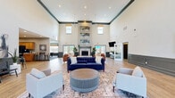 Located at 4007 Raynor Pkwy in Bellevue, Nebraska, this well-appointed single-floor community space offers an impressive blend of comfort and functionality. The centerpiece is a soaring living room with dramatic double-height ceilings, elegant wainscoting in a dark gray finish, and light wood-look flooring that runs throughout. Multiple seating arrangements — including rich leather sofas, accent chairs, and area rugs — create distinct gathering zones. A classic fireplace anchors one wall, while large windows and glass French doors flood the space with natural light and provide views of an outdoor pool area. A ceiling fan adds a finishing touch overhead. The kitchen is generously sized, featuring warm honey-toned cabinetry, white countertops, a sage green subway tile backsplash, and black appliances including a built-in microwave. Bar-height tables with industrial-style stools offer casual dining options. The game room, with plush patterned carpeting and coordinating wainscoting, houses a full-size billiard table, a wall-mounted television, and high-top seating, making it an ideal recreational retreat. Throughout, the space maintains a cohesive, modern-transitional aesthetic with curated artwork and thoughtful design details.