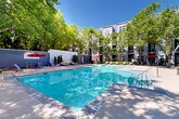 Located at 2115 Piedmont Rd NE in Atlanta's vibrant Buckhead/Piedmont corridor, this outdoor amenity space offers a well-appointed resort-style experience set within a lushly landscaped courtyard. The centerpiece is a generously sized swimming pool with crystal-clear blue water, featuring a gradual entry and defined depth markings — ideal for both relaxing and recreational use. The pool deck is finished in smooth concrete, providing ample space for sunbathing and casual lounging.

Surrounding the pool, mature deciduous trees create a natural canopy that offers shade and a sense of seclusion, softening the urban environment and lending the space a park-like ambiance. Red-accented market umbrellas are positioned throughout, shading clusters of lounge chairs, chaise lounges, and outdoor dining tables that invite both social gatherings and quiet relaxation.

A decorative feature wall with vertical plantings serves as a polished architectural backdrop at one end of the pool, adding visual depth and design intentionality to the space. A secondary courtyard area adjacent to the pool provides additional seating and gathering space beneath the tree canopy, enclosed by iron fencing and bordered by manicured shrubs and seasonal plantings. The multi-story residential buildings surrounding the courtyard feature a contemporary grey exterior, framing the outdoor space with a cohesive modern aesthetic.