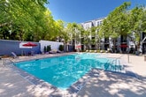 Located at 2115 Piedmont Rd NE in Atlanta's vibrant Buckhead/Piedmont corridor, this outdoor amenity space offers a well-appointed resort-style experience set within a lushly landscaped courtyard. The centerpiece is a generously sized swimming pool with crystal-clear blue water, featuring a gradual entry and defined depth markings — ideal for both relaxing and recreational use. The pool deck is finished in smooth concrete, providing ample space for sunbathing and casual lounging.

Surrounding the pool, mature deciduous trees create a natural canopy that offers shade and a sense of seclusion, softening the urban environment and lending the space a park-like ambiance. Red-accented market umbrellas are positioned throughout, shading clusters of lounge chairs, chaise lounges, and outdoor dining tables that invite both social gatherings and quiet relaxation.

A decorative feature wall with vertical plantings serves as a polished architectural backdrop at one end of the pool, adding visual depth and design intentionality to the space. A secondary courtyard area adjacent to the pool provides additional seating and gathering space beneath the tree canopy, enclosed by iron fencing and bordered by manicured shrubs and seasonal plantings. The multi-story residential buildings surrounding the courtyard feature a contemporary grey exterior, framing the outdoor space with a cohesive modern aesthetic.
