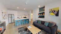Located at 1006 SE 3rd St in Bentonville, Arkansas, this single-floor residence offers a thoughtfully designed open-concept layout with contemporary finishes throughout. The living and kitchen areas flow seamlessly together, anchored by polished concrete floors that lend an industrial-modern character to the space. The kitchen is outfitted with sleek white flat-panel cabinetry, a full suite of stainless steel appliances including a refrigerator, dishwasher, and electric range, and a striking teal island with a white quartz countertop and bar seating — a bold design accent that defines the space. The living area is warm and inviting, featuring built-in teal-accented shelving, a wall-mounted media console, and large windows and a glass door that draw in generous natural light and provide outdoor views. The bedroom is a serene retreat with bright white walls, natural light from a well-positioned window, and ample room for comfortable furnishings. The bathroom maintains the home's clean aesthetic with a white vanity, large mirror, and a tub-shower combination. Throughout the residence, a cohesive palette of white, teal, and neutral tones creates a modern, refreshing ambiance that is both functional and visually appealing.