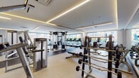 This contemporary fitness center is thoughtfully designed to support a full range of workout routines across multiple dedicated areas. The main gym floor is expansive and bright, bathed in natural light from large floor-to-ceiling windows that offer outdoor views. Recessed linear LED lighting along the ceiling adds a polished, modern ambiance throughout. The space is outfitted with a comprehensive selection of Life Fitness equipment, including a row of treadmills, elliptical machines, stationary bikes, cable and pulley strength machines, benches, and a well-stocked free weights area with dumbbells and barbells on organized racks. Large wall mirrors throughout the main floor enhance the sense of openness and allow for form monitoring during workouts. A separate cycling studio features vivid blue accent walls, floor-to-ceiling mirrors, and a fleet of Life Cycle spin bikes, creating an immersive and energizing environment for group or solo cycling sessions. An additional functional training area provides stability balls, exercise mats, and medicine balls for stretching and accessory work. Storage cubbies and thoughtfully placed flooring transitions between rubber and carpet zones further reflect the facility's attention to detail and user experience.