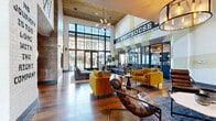 The Westhouse Clubhouse at 11325 Park Vista Blvd is a generously sized, single-story community gathering space that masterfully blends industrial and modern design elements. The main lounge is immediately striking, boasting double-height ceilings clad in warm wood planking, floor-to-ceiling black-framed windows that flood the interior with natural light, and a dramatic large-scale "Westhouse" mural that anchors the space with bold community identity. Rich hardwood floors, a statement chandelier, and an eclectic mix of leather, mustard-yellow, and navy upholstered seating create an inviting yet energetic atmosphere.

A central tiled fireplace column divides the space into distinct lounge areas, while vibrant Western-themed murals and rotating community event boards contribute to the clubhouse's lively character. A dedicated café-style nook features high-top tables with red metal stools, a coffee station, and mounted flat-screen displays. The fully equipped kitchen area showcases sleek dark cabinetry with brushed gold hardware, stainless steel appliances, a wall oven, and a large island with bar seating beneath globe pendant lights.

A bright conference and dining area with polished concrete floors and full-width sliding glass doors opens to stunning views of the pool courtyard and surrounding community. A business center with workstations rounds out the functional offerings of this well-appointed clubhouse.