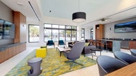 This single-floor communal lounge is a thoughtfully designed social space that blends contemporary architecture with vibrant, eclectic interiors. The space is anchored by soaring ceilings and expansive floor-to-ceiling windows that flood the interior with natural light while framing views of a resort-style pool lined with palm trees, as well as panoramic city vistas.

The main lounge area features large-format light tile flooring and a bold, patterned area rug in yellow and teal tones, setting the foundation for an eclectic furniture arrangement. Seating includes a substantial dark leather sofa, swivel lounge chairs in muted grays and teals, and colorful accent ottomans in yellow and green. A wall-mounted flat-screen television is set against a textured gray panel flanked by warm wood cabinetry, creating a focal media wall.

A dedicated dining nook runs alongside the window wall, featuring a long solid wood table paired with dark chairs and a continuous teal upholstered banquette bench, illuminated by a trio of pendant lights. Adjacent to the entry is a sleek wet bar with dark countertops, warm wood cabinetry, and bar seating. A built-in shelving unit adds architectural detail near the entrance. The overall ambiance is modern, energetic, and welcoming.