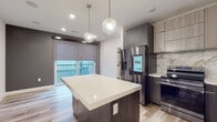Located at 5655 Pershing Ave in St. Louis, Missouri, this contemporary single-floor residence showcases a thoughtfully designed layout with high-end finishes throughout. The open-concept kitchen is a true centerpiece, featuring dark wood cabinetry paired with lighter upper cabinets, a striking marble-look backsplash, white quartz countertops, and a spacious central island with pendant globe lighting overhead. Stainless steel appliances, including a smart refrigerator and electric range, complete the chef-inspired space. Light wood-tone flooring flows seamlessly throughout the unit, adding warmth and continuity to the modern aesthetic. The living area benefits from large windows fitted with roller shades, offering direct access to a private balcony and allowing an abundance of natural light to fill the space. The bathroom is elegantly appointed with hexagonal dark tile flooring, a floating wood-grain vanity with matte black hardware, a bold framed mirror with statement lighting, and a marble-accented shower enclosure. A stackable laundry unit is also integrated into the layout for added convenience. Clean lines, neutral tones, and carefully selected fixtures give the space a cohesive, upscale feel throughout.