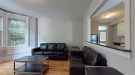 This single-floor unit offers a well-organized layout with distinct living, sleeping, and functional zones throughout. The living area is furnished with dark leather sofas and low-profile coffee tables set atop warm hardwood floors, creating a comfortable common space. A pass-through opening connects the living room to an adjacent area, adding an open, connected feel to the interior. Natural light filters in through multiple windows, brightening the neutral gray and white walls throughout.

The kitchen features stainless steel appliances, white cabinetry, and hardwood flooring, with a dedicated exterior door providing convenient direct access. The bedrooms are carpeted and include built-in or freestanding storage, with several rooms offering windows that overlook greenery outside. One bedroom benefits from a bay-style windowed alcove, adding character and additional natural light.

Two bathrooms serve the unit — one featuring tile flooring, a pedestal sink, and a soaking tub, while the second is a more compact half-bath with tile floors and clean white fixtures. Wainscoting detailing and clean architectural moldings appear in select rooms, lending a classic touch. The overall ambiance is practical, bright, and functional, suited for multi-occupant living.