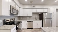 This efficiently designed single-floor space makes excellent use of its footprint, offering a bright and modern interior with a clean, contemporary aesthetic throughout. The kitchen is the standout feature, showcasing white shaker-style cabinetry extending to the ceiling, granite-look countertops, a subway tile backsplash, and a full suite of stainless steel appliances including a range, microwave, dishwasher, and refrigerator. A central kitchen island with matching countertop and built-in storage provides additional prep space and cabinetry. Wood-look luxury vinyl plank flooring runs throughout, adding warmth and cohesion to the open layout. Recessed lighting illuminates the main living and kitchen areas, contributing to a bright and inviting atmosphere. The space includes two bathrooms, one of which features a soaking tub with tiled surround, a granite-topped vanity with white cabinetry, and wood-look flooring. A dedicated closet conveniently houses a stacked washer and dryer alongside wire shelving for storage. The overall design is characterized by a neutral color palette of whites and soft grays, creating a fresh, move-in-ready environment that feels clean and well-finished.
