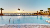 Located along the waterfront in Parker, Florida, this resort-style pool area offers a beautifully designed outdoor retreat with breathtaking views of the bay and a neighboring bridge. The expansive swimming pool features elegant paver decking and is framed by towering palm trees that enhance the coastal atmosphere. Surrounding the pool, a generous sunbathing deck is outfitted with rows of chaise lounges and shaded seating areas with umbrellas, creating an ideal environment for relaxation under the Florida sun.

Adjacent to the pool, a covered outdoor dining pavilion provides a refined al fresco experience. Large sliding glass panels open the space to panoramic waterfront vistas, while ceiling fans maintain comfortable airflow throughout. A long communal dining table with ample seating makes this pavilion well-suited for both casual gatherings and organized events. Recessed lighting and clean architectural finishes give the pavilion a polished, contemporary feel.

The surrounding grounds feature well-maintained landscaping with tropical plantings and wide concrete pathways connecting the pool area to the broader residential community. The backdrop of multi-story residential buildings clad in coastal siding complements the overall aesthetic. Whether at sunrise or sunset, the waterfront setting delivers a consistently striking and serene ambiance throughout this thoughtfully designed outdoor space.
