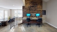 This single-floor business center combines rustic character with functional design, creating a welcoming and productive environment. The space is organized across two distinct zones connected by a split-level layout with an iron railing staircase that adds an architectural focal point.

The workstation area is defined by a dramatic reclaimed wood accent wall, providing a warm, textured backdrop for a granite-topped desk accommodating two computer workstations. A large decorative iron clock mounted on the wood wall adds a distinctive design element, reinforcing the rustic-industrial aesthetic. Natural light streams in through white-framed windows, brightening the workspace.

The adjacent meeting or collaborative area features a substantial reclaimed wood table surrounded by modern blue chairs, ideal for group gatherings or informal meetings. A charming bay window with a built-in wood seat bench floods this area with additional natural light and offers views of the surrounding outdoor greenery. The neutral taupe walls, white trim, and carpeted flooring throughout contribute to a calm, professional atmosphere that balances comfort with practicality.