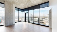 Located on a high floor at 81 E 14th St in the heart of Kansas City, MO, this contemporary unit delivers a striking urban living experience defined by its industrial-modern aesthetic. The open living area is anchored by expansive corner floor-to-ceiling glass walls framed in sleek black steel, flooding the space with natural light and offering breathtaking panoramic views of the Kansas City skyline and surrounding cityscape. Exposed concrete ceilings lend an industrial character throughout, complemented by light hardwood flooring that adds warmth and continuity across the space.

The modern kitchen is thoughtfully designed with a full suite of stainless steel appliances, including a French door refrigerator, electric range, built-in microwave, and dishwasher. Cabinetry in a two-tone finish of white gloss and natural wood tones lines the walls, paired with white quartz countertops and a marble-look tile backsplash. Exposed ductwork on the ceiling reinforces the loft-inspired character.

The bathrooms feature large-format marble-look tile flooring, white quartz vanity tops, wood-framed floating vanities, and marble-tiled shower surrounds. A private balcony accessible through the living area extends the experience outdoors, offering unobstructed views across the Kansas City urban landscape in multiple directions.
