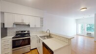 Located at 7550 N Lombard St in Portland's St. Johns neighborhood, Unit 303 is a single-floor residence designed with a clean, modern aesthetic. Warm hardwood-style flooring runs throughout the living areas and bedroom, creating a cohesive and inviting atmosphere. The open-concept layout connects the living area to the kitchen, which features white shaker cabinetry, open shelving, quartz countertops, a subway tile backsplash, stainless steel appliances — including a gas range and dishwasher — and a peninsula with a built-in sink that doubles as a casual dining or prep space. The living area is bright and open, with a door that leads to a view of surrounding greenery, filling the space with natural light. The bedroom is a comfortable, well-proportioned room with a window overlooking mature trees, offering a serene outdoor view. A closet with louvered bi-fold doors provides ample storage. The bathroom includes a full soaking tub and shower combination with clean white surrounds and chrome fixtures. A stacked washer and dryer unit is conveniently housed within the unit, adding to the overall practicality of the space. The layout is efficient and functional, making effective use of every area.
