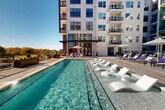 Located at 2408 South Blvd in Charlotte's vibrant South End neighborhood, this outdoor amenity deck offers a thoughtfully designed resort-style experience. The centerpiece is a generously sized swimming pool with crystal-clear water, complemented by in-water lounge chairs that allow residents to relax while partially submerged. Surrounding the pool is a spacious sun deck finished in neutral-toned pavers, lined with an abundance of purple and white chaise lounges, striped market umbrellas, and sleek side tables, creating a lively yet sophisticated atmosphere. A pergola structure provides a shaded gathering area, enhancing the deck's versatility for both relaxation and socializing. The space is framed by the multi-story modern residential building, characterized by its white exterior, large windows, and private balconies visible above. A landscaped terrace area features raised planters filled with colorful seasonal flowers, ornamental grasses, and sculptured shrubs, bordered by glass-panel railings that open up to sweeping views of the Charlotte skyline and surrounding treetops. The open-sky setting ensures abundant natural light throughout the day, making this outdoor amenity deck a standout feature of the property.