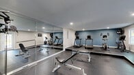 Located at 10100 Brown Bridge Road in Covington, GA, this dedicated exercise room offers a comprehensive fitness facility within a single, thoughtfully designed space. The room features durable rubber flooring throughout, providing both safety and functionality for a wide range of workout activities.

The cardio zone is anchored by two treadmills and an elliptical machine, accompanied by a recumbent bike, all positioned along a striking blue accent wall. Large industrial fans are mounted on the wall to ensure proper airflow during intense workouts. A multi-functional cable machine serves as the centerpiece of the strength training area, complemented by a weight bench and free weights. Wall-mounted yoga mats and stability balls provide options for stretching and core training.

A large mirror spans one wall, enhancing the sense of space and allowing users to monitor their form. A wall-mounted flat-screen TV adds an entertainment element to the workout experience. Recessed lighting illuminates the room evenly, while a window allows natural light to filter in. A wall-mounted handrail along one side adds a practical safety feature. The clean, neutral color palette of white walls contrasted with the bold blue accent wall creates a focused, energizing atmosphere ideal for fitness use.