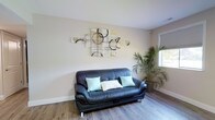 This single-floor space offers a clean, modern aesthetic defined by warm neutral wall tones and durable hardwood-style flooring that runs throughout. The living area serves as the heart of the space, featuring a sleek black leather sofa accented with soft mint and white cushions, creating a comfortable and inviting atmosphere. A striking multi-panel abstract wall art piece commands attention above the seating area, adding a creative focal point with its blend of earthy and golden tones. Natural light filters in through a window, brightening the room and highlighting the airy, open feel of the interior. A lush indoor palm plant positioned near the window introduces a touch of nature, enhancing the calm and refreshing ambiance. The space is finished with clean white baseboards and trim, contributing to a crisp, polished look. Subtle security features, including a ceiling-mounted camera, suggest the space may serve a professional or semi-public function. Overall, the layout and design reflect a thoughtfully arranged, versatile environment suitable for relaxation or reception purposes.