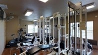 This fitness center offers a dedicated space for strength training and conditioning, thoughtfully outfitted with a range of professional-grade equipment. The room features multiple cable machines, including Body Masters multi-station units, along with adjustable benches and weight systems that cater to a variety of exercise routines. The layout is organized to allow users to move efficiently between stations, maximizing the functionality of the available space. Large windows along the walls allow natural light to filter into the room, creating a bright and energizing workout environment. The textured ceiling is fitted with recessed fluorescent lighting panels, ensuring consistent illumination throughout. Wall-mounted mirrors reflect the equipment and amplify the sense of openness within the space. A stepladder and stability ball visible in the background suggest additional versatile training options. The flooring provides a sturdy, supportive surface suitable for a fitness setting. Overall, this fitness center is well-suited for users seeking a comprehensive strength training facility, combining practical equipment arrangement with a clean, functional aesthetic.