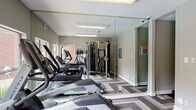 This fitness center is a compact, single-floor facility designed to support a well-rounded workout routine. The interior is finished with a striking chevron-patterned vinyl floor in shades of grey and white, adding a modern, dynamic feel to the space. Neutral grey walls and recessed fluorescent lighting create a clean, functional atmosphere throughout.

The cardio zone is well-appointed with multiple treadmills, an elliptical trainer, and a stationary spin bike, all positioned along one wall to maximize floor space. A multi-station cable machine provides comprehensive strength training options, rounding out the equipment selection. Large floor-to-ceiling mirrors line one wall, enhancing the sense of space and allowing users to monitor their form during exercise.

Natural light fills the room through several windows that look directly out onto the adjoining outdoor pool area, offering a pleasant view during workouts. A glass-paneled door provides direct access to the pool deck, which features a generously sized swimming pool surrounded by a spacious sun-soaked patio. The pool area is furnished with blue lounge chairs, umbrella-shaded tables, and is bordered by mature trees and flowering shrubs, creating a serene and inviting outdoor retreat. The exterior of the building features classic brick construction, complementing the surrounding residential architecture.