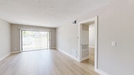 This single-floor space presents a clean, contemporary aesthetic throughout, defined by light hardwood-style flooring, neutral gray walls, and white trim that create a cohesive and airy feel. The open living and dining area is generously proportioned, anchored by a modern dual-ring pendant light fixture and a full-width sliding glass door fitted with vertical blinds, allowing natural light to flow in while offering access to the outdoors. Textured ceilings are present throughout, and HVAC vents are consistently integrated into both the walls and floors.

The kitchen is designed with functionality and style in mind, featuring white shaker-style cabinetry that extends to the upper walls, light quartz countertops, a deep single-basin stainless steel sink with a flexible coil-neck faucet, and a stainless steel dishwasher. The layout offers a practical L-shaped workspace with ample storage.

Additional rooms include a well-sized bedroom with a window fitted with horizontal blinds, providing natural light and privacy. A separate room with built-in ventilation panels is also accessible from the main living area. The overall ambiance is bright, clean, and move-in ready.