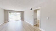 This single-floor space presents a clean, contemporary aesthetic throughout, defined by light hardwood-style flooring, neutral gray walls, and white trim that create a cohesive and airy feel. The open living and dining area is generously proportioned, anchored by a modern dual-ring pendant light fixture and a full-width sliding glass door fitted with vertical blinds, allowing natural light to flow in while offering access to the outdoors. Textured ceilings are present throughout, and HVAC vents are consistently integrated into both the walls and floors.

The kitchen is designed with functionality and style in mind, featuring white shaker-style cabinetry that extends to the upper walls, light quartz countertops, a deep single-basin stainless steel sink with a flexible coil-neck faucet, and a stainless steel dishwasher. The layout offers a practical L-shaped workspace with ample storage.

Additional rooms include a well-sized bedroom with a window fitted with horizontal blinds, providing natural light and privacy. A separate room with built-in ventilation panels is also accessible from the main living area. The overall ambiance is bright, clean, and move-in ready.