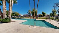 Located at 1106 W Bell Rd in Phoenix, Arizona, this single-level outdoor amenity area serves as a central gathering space within a residential community. The centerpiece is a generously sized swimming pool with clear, blue-green water, complemented by a separate spa, both accessible via stainless steel handrails and surrounded by a wide, paved pool deck. Lounge chairs are positioned throughout the deck, offering ample space for sunbathing and relaxation beneath the abundant Arizona sunshine. Towering palm trees and mature shade trees are strategically placed around the perimeter, providing natural relief from the desert heat while lending a resort-like atmosphere to the setting. A well-maintained lawn area stretches beyond the pool enclosure, bordered by decorative iron fencing that defines the space. Desert-landscaped rock beds with low-water plantings frame the walkways leading to and from the pool area. A single-story community building with a tile roof and stucco exterior sits adjacent to the pool, likely housing restrooms or amenity facilities. Patio tables and chairs are available for outdoor dining and socializing. The overall environment reflects the classic Arizona outdoor lifestyle, combining functionality with a relaxed, sun-soaked aesthetic.