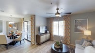 This single-floor residence offers a welcoming and well-appointed living area designed with both comfort and style in mind. The main living space is anchored by a sleek, modern ceiling fan with an integrated light fixture, ensuring year-round comfort and ambient lighting. Large windows dressed in vibrant, patterned curtains in warm tones of orange, gold, and gray allow generous amounts of natural light to flood the room, creating a bright and airy atmosphere. A sliding glass door provides direct access to an outdoor patio area, seamlessly connecting the interior to the exterior. The living area features light wood-look flooring that enhances the open feel of the space, complemented by a textured area rug. A mid-century modern media console serves as a focal point, providing both storage and a surface for the mounted flat-screen television. The room is accented with tasteful décor, including a round dark-toned coffee table adorned with a small potted succulent arrangement, adding a touch of natural greenery. The neutral gray walls and textured white ceiling provide a clean, versatile backdrop that ties the entire space together, making it both functional and visually appealing.