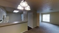 This single-story space showcases an open-concept layout that connects the kitchen and living areas seamlessly. A raised breakfast bar serves as a natural dividing element between the two zones, topped with a light countertop surface and illuminated by a multi-bulb overhead light fixture that provides ample task lighting. The kitchen area features a skylight, enhancing natural brightness, while the adjacent living space benefits from a window that allows additional daylight to filter in. Flooring transitions from dark hardwood-style material in the kitchen and hallway to plush carpeting in the living area, creating a clear distinction between functional and relaxation zones. The textured popcorn ceiling adds a tactile dimension overhead throughout the space. A central hallway visible from the main area leads to additional rooms, with a door framed by neutral-toned walls. The overall color palette consists of soft grays and beiges, contributing to a calm, neutral ambiance. The layout feels practical and well-connected, with each area flowing naturally into the next.