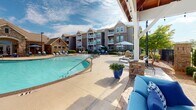 Located at 3434 Laurens Rd in Greenville, South Carolina, this outdoor amenity space offers a resort-style experience centered around a generously sized, freeform swimming pool with a smooth, light-colored concrete deck. The pool area is outfitted with multiple lounge chairs and market umbrellas, providing ample space for relaxation under the South Carolina sun.

A standout feature of the space is the covered pavilion, which boasts soaring vaulted ceilings with warm wood planking and exposed timber framing. Beneath the pavilion, a striking floor-to-ceiling stacked stone fireplace anchors the seating area and supports a mounted outdoor television, creating an ideal setting for year-round outdoor entertaining. Comfortable blue lounge furniture and a paver brick floor complete the pavilion's inviting atmosphere.

The perimeter of the pool area is defined by wrought iron fencing with stone pillar accents, complemented by neatly manicured shrubs and mature trees that provide both privacy and shade. Decorative potted flowering plants add vibrant color throughout the space. The surrounding multi-story residential buildings feature a blend of stone and neutral siding, with private balconies overlooking the amenity area. The overall ambiance is polished, welcoming, and well-appointed, reflecting a high standard of outdoor community living.