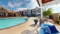 Located at 3434 Laurens Rd in Greenville, South Carolina, this outdoor amenity space offers a resort-style experience centered around a generously sized, freeform swimming pool with a smooth, light-colored concrete deck. The pool area is outfitted with multiple lounge chairs and market umbrellas, providing ample space for relaxation under the South Carolina sun.

A standout feature of the space is the covered pavilion, which boasts soaring vaulted ceilings with warm wood planking and exposed timber framing. Beneath the pavilion, a striking floor-to-ceiling stacked stone fireplace anchors the seating area and supports a mounted outdoor television, creating an ideal setting for year-round outdoor entertaining. Comfortable blue lounge furniture and a paver brick floor complete the pavilion's inviting atmosphere.

The perimeter of the pool area is defined by wrought iron fencing with stone pillar accents, complemented by neatly manicured shrubs and mature trees that provide both privacy and shade. Decorative potted flowering plants add vibrant color throughout the space. The surrounding multi-story residential buildings feature a blend of stone and neutral siding, with private balconies overlooking the amenity area. The overall ambiance is polished, welcoming, and well-appointed, reflecting a high standard of outdoor community living.