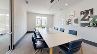Located at 3000 Cypress Lake Rd in North Charleston, South Carolina, this single-floor space is designed as a contemporary coworking and community amenity environment. The layout is thoughtfully organized into distinct functional zones, each serving a different style of work or relaxation.

The main lounge area features high-back teal booth seating with integrated side tables, ideal for focused individual work or semi-private conversations. Complementing these booths are sculptural cream lounge chairs arranged around dark pedestal tables, creating a casual yet polished gathering area. Patterned carpet tiles in neutral tones unify the space underfoot, while modern ring pendant light fixtures provide warm, directed illumination overhead.

Large windows fitted with roller shades line multiple walls, allowing generous natural light to filter throughout while maintaining glare control. A sunlit café-style nook offers small round tables paired with woven-seat bistro chairs, positioned directly beside floor-to-ceiling windows with outdoor views of the surrounding landscape.

A dedicated conference room accommodates a full boardroom-style table surrounded by navy blue executive chairs, lit by a sleek linear pendant fixture and accented with abstract wall art. The overall aesthetic is clean and contemporary, with white walls, recessed lighting, and a cohesive color palette of teal, cream, and navy throughout.