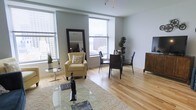 This single-floor space showcases a seamlessly integrated living and dining area defined by light hardwood flooring that runs throughout, lending warmth and continuity to the open-plan layout. Large windows line multiple walls, flooding the interior with abundant natural light while framing impressive elevated views of the surrounding urban skyline. The living area features a plush area rug anchoring a glass coffee table and a classic accent chair, complemented by a full-length mirror that enhances the sense of space. A pendant light fixture adds a contemporary touch overhead. The dining area is furnished with a sleek dark table and matching chairs, positioned to take full advantage of the corner windows and city views beyond. Soft grey-toned walls provide a neutral backdrop that pairs well with the natural wood floors, creating a clean and modern aesthetic throughout. The overall ambiance is bright, airy, and sophisticated, well-suited for both relaxed everyday living and casual entertaining.