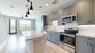 This two-story residence offers a well-designed layout divided equally across two levels, combining modern finishes with functional living spaces. The open-concept kitchen and living area anchors the main floor, showcasing grey shaker-style cabinetry, granite countertop surfaces, and a geometric white tile backsplash. A central island with matching granite provides additional workspace and casual dining potential, while sleek black pendant lights hang overhead. Stainless steel appliances, including a range, microwave, and dishwasher, complete the kitchen. Light-toned hardwood flooring runs throughout the main living areas, complementing the neutral grey wall palette and allowing natural light from the windows to brighten the space.

The bedroom is carpeted and comfortably sized, featuring a window with outdoor views, a dedicated closet, and a dark five-panel door that adds a contrasting design detail. Two bathrooms serve the residence; one features grey cabinetry, a granite-topped vanity, a framed mirror, and light hardwood flooring. A hallway and closet provide additional storage and circulation. The overall aesthetic is clean and contemporary, with a cohesive grey-and-neutral color scheme carried consistently throughout both floors.