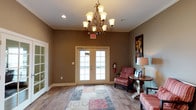 This single-floor space makes a strong first impression with its thoughtfully designed entryway and lobby area. The entrance is anchored by a striking ornate wrought-iron chandelier with frosted glass shades, casting warm, ambient light across the room. Neutral warm-taupe walls are accented by crisp white crown molding, lending the space a polished and refined character.

Natural light flows generously through a set of classic white-framed French doors, which serve as the main entry point and connect the interior to the outdoors. The flooring throughout features rich hardwood-style planks, grounded by a decorative area rug in muted red and gray tones that adds texture and visual warmth.

A comfortable seating arrangement near the entrance includes upholstered striped armchairs and a classic wooden side table topped with a traditional lamp, creating an inviting waiting or reception area. Decorative wall art adds a touch of personality to the space.

Recessed lighting complements the chandelier to ensure the space is well-lit throughout. Overall, the design balances comfort and functionality, making it well-suited as a welcoming reception or common area for visitors and occupants alike.