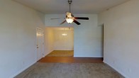This single-story space features an open-concept layout connecting a living area to a dining space, creating a generous, flowing floor plan. The living area is distinguished by vaulted ceilings, which lend a sense of openness and volume to the room. A large ceiling fan with a built-in light fixture is centrally mounted, providing both air circulation and ambient lighting. The flooring in the living area consists of neutral-toned carpet, offering a soft underfoot feel.

A wide, open passageway leads into the adjoining dining area, which continues the light, airy aesthetic of the space. This secondary area features its own elegant chandelier, adding a refined touch to the room. The flooring transitions to hard surface material in this zone, providing a practical distinction between the two spaces.

The walls throughout are painted in a soft, neutral tone, contributing to a clean and understated aesthetic. A paneled entry door is positioned along one wall, providing access to the exterior. White baseboards frame the perimeter of each room, adding a crisp finishing detail. Overall, the space presents a functional and straightforward layout with comfortable proportions throughout.