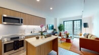This single-floor residence showcases a thoughtfully designed open-concept layout with a seamless flow between the kitchen, dining, and living areas. The kitchen is outfitted with flat-front wood-grain cabinetry extending to the ceiling, under-cabinet LED lighting, white quartz countertops, a central island with additional storage, and a full suite of stainless steel appliances including a French door refrigerator, range, microwave, and dishwasher. Light wood-tone flooring runs throughout, reinforcing a cohesive, contemporary aesthetic.

The living area is anchored by floor-to-ceiling sliding glass doors that frame expansive urban views, filling the space with abundant natural light. The bedroom continues this connection to the outdoors with its own large windows offering cityscape views, while light wood flooring and clean white walls maintain a calm, airy atmosphere.

The bathroom features a floating wood-grain vanity with a white quartz countertop, an illuminated wide-format mirror, tile flooring, and a full soaking tub with a tiled surround. Recessed lighting throughout the residence ensures consistent, well-distributed illumination, and the neutral palette across all rooms contributes to a polished, modern ambiance.