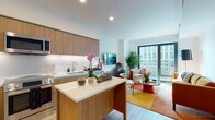 This single-floor residence showcases a thoughtfully designed open-concept layout with a seamless flow between the kitchen, dining, and living areas. The kitchen is outfitted with flat-front wood-grain cabinetry extending to the ceiling, under-cabinet LED lighting, white quartz countertops, a central island with additional storage, and a full suite of stainless steel appliances including a French door refrigerator, range, microwave, and dishwasher. Light wood-tone flooring runs throughout, reinforcing a cohesive, contemporary aesthetic.

The living area is anchored by floor-to-ceiling sliding glass doors that frame expansive urban views, filling the space with abundant natural light. The bedroom continues this connection to the outdoors with its own large windows offering cityscape views, while light wood flooring and clean white walls maintain a calm, airy atmosphere.

The bathroom features a floating wood-grain vanity with a white quartz countertop, an illuminated wide-format mirror, tile flooring, and a full soaking tub with a tiled surround. Recessed lighting throughout the residence ensures consistent, well-distributed illumination, and the neutral palette across all rooms contributes to a polished, modern ambiance.