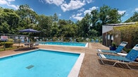 This outdoor aquatic facility features two separate swimming pools set within a spacious, tree-lined environment. The larger pool is equipped with lap lane dividers, making it suitable for both recreational swimming and structured exercise, while the smaller pool provides a more intimate setting for leisurely use. Both pools display clear, vivid blue water and are framed by wide concrete and aggregate sundecks furnished with an abundance of blue lounge chairs and shaded seating areas complete with umbrella-covered tables. The entire complex is enclosed by a chain-link fence, ensuring safety and privacy, while lush mature trees — including tall pines and dense deciduous growth — create a natural green backdrop that lends a tranquil, secluded atmosphere. Potted plants and landscaped shrubs add decorative touches throughout the deck areas. A small utility or pool house structure is positioned adjacent to the pools, providing operational support for the facility. The space benefits from ample open sky, allowing for abundant natural sunlight across the decks. Overall, the facility offers a well-maintained, comfortable outdoor retreat designed to accommodate a variety of aquatic and leisure activities.
