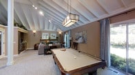 This single-floor space is thoughtfully designed as a combined recreation and lounge area, anchored by a full-size pool table positioned beneath a striking geometric pendant light fixture. The vaulted ceiling is one of the room's most prominent architectural features, showcasing exposed white painted rafters that lend the space an open, airy quality and a sense of generous vertical height.

The walls are finished in a warm taupe tone that complements the neutral carpet flooring, creating a cohesive and inviting atmosphere throughout. Track lighting runs along the ceiling, providing flexible illumination alongside wall-mounted sconces that add a softer ambient glow.

Adjacent to the game area, a comfortable seating arrangement with upholstered sofas and accent furniture forms a relaxed lounge zone, enhanced by decorative artwork and a fireplace visible in the background. Patio doors allow natural light to filter into the space, offering a connection to the outdoors.

Overall, the space balances recreational function with refined interior design, making it well-suited for entertaining guests or unwinding in a stylish setting.